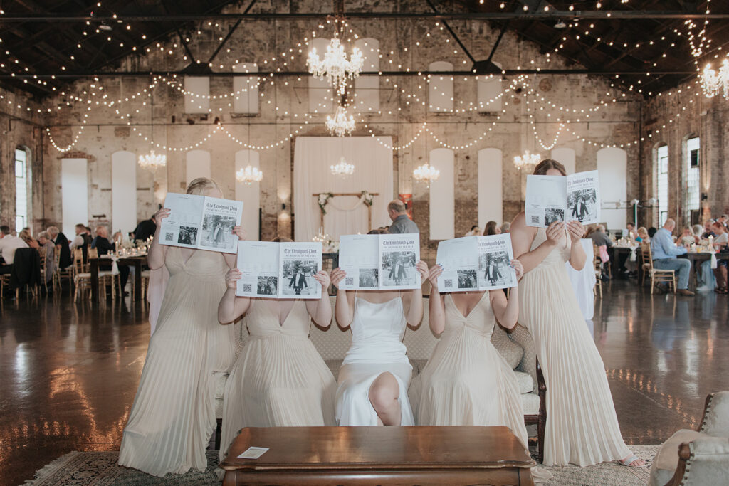 Bride and bridesmaids hold the wedding newspapers in front of their faces