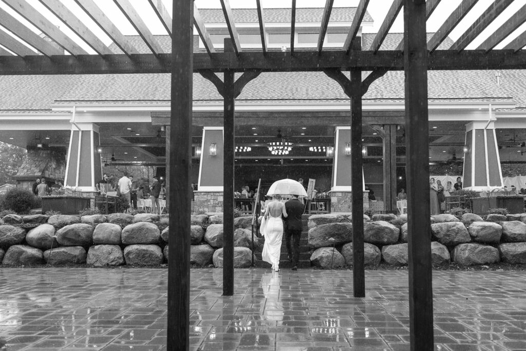 Bride and groom walk up the steps to the pavillion as rain falls on their shared clear umbrella.